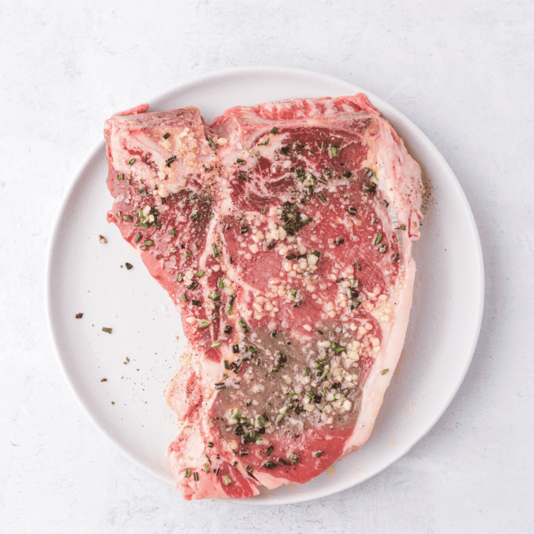Seasoning a T-bone steak with olive oil, salt, pepper, rosemary, and minced garlicl letting it rest at room temperature.