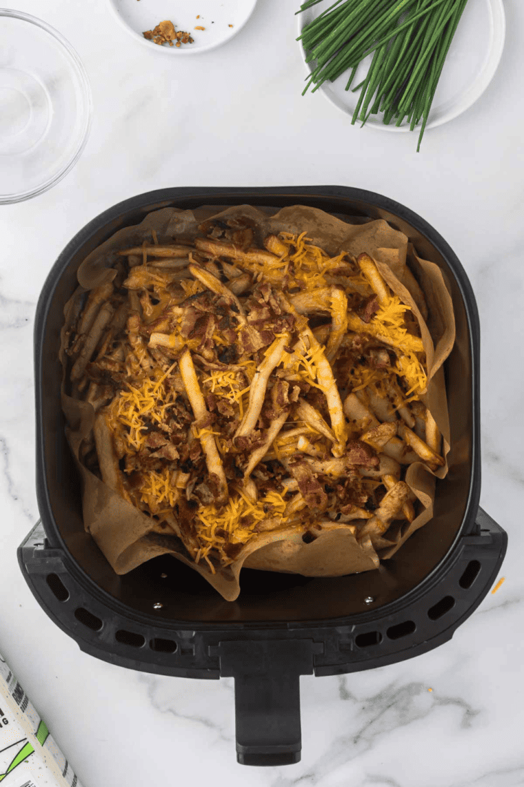 Seasoned French fries in a heat-safe dish, topped with shredded cheddar, Monterey Jack cheese, and crumbled bacon, ready to melt in the air fryer.