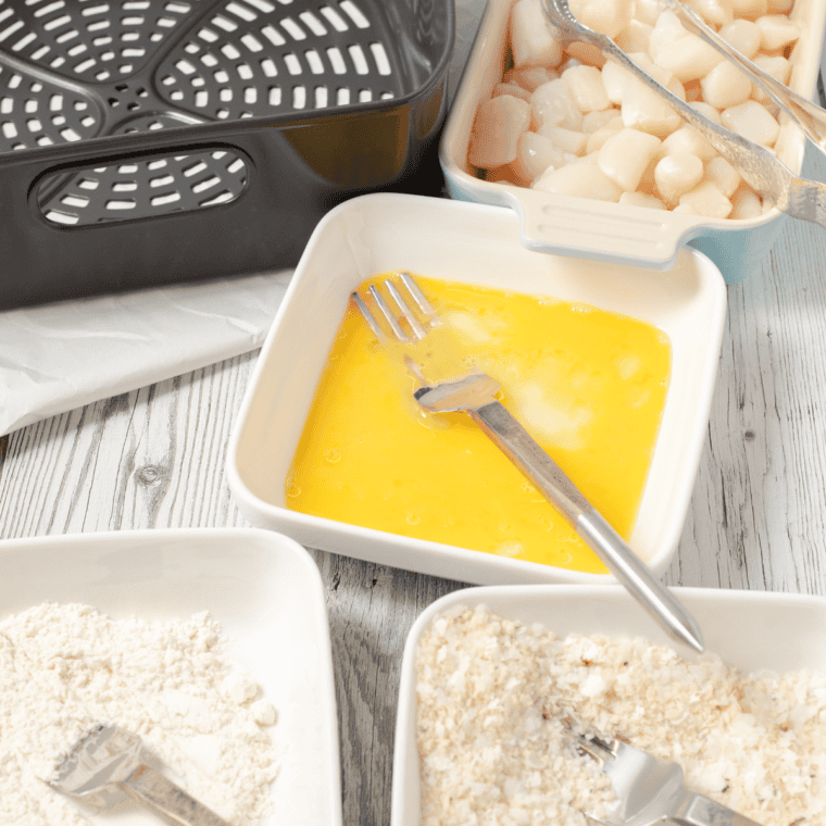 Three shallow bowls filled with flour, beaten eggs, and seasoned panko mixture for breading scallops.