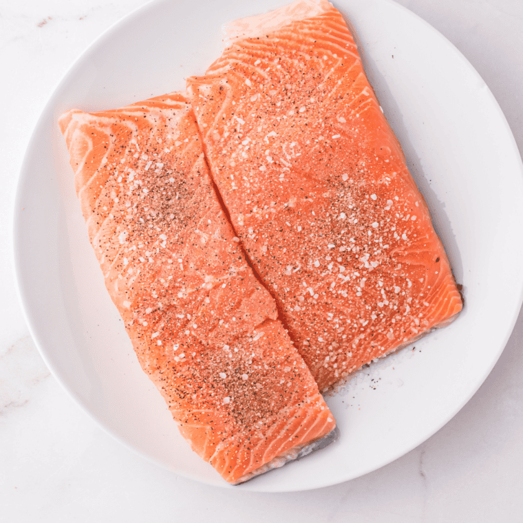 Salmon fillets being patted dry with a paper towel and seasoned with salt and pepper on white plate.