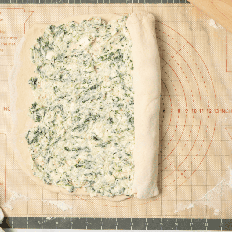 Spreading spinach, garlic butter, and mozzarella mixture on rolled-out pizza dough, then rolling and slicing into spiraled rounds