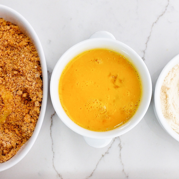 Three bowls lined up for dredging: one with coconut flour and salt, one with beaten eggs, and one with seasoned pork rind coating.