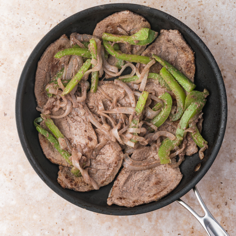 Steak browning in a skillet with sautéed onions and peppers until tender.
