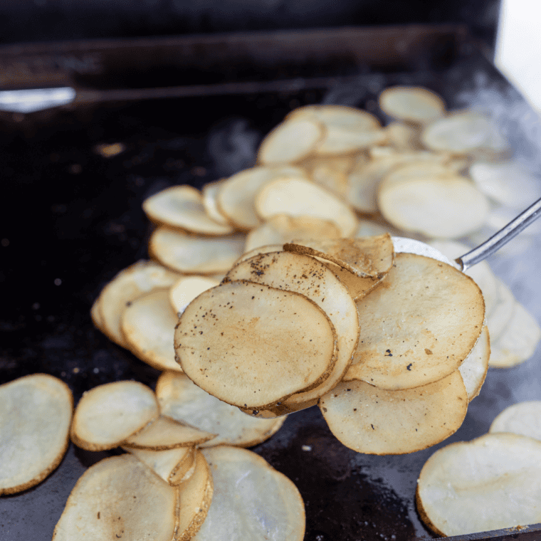Thin potato slices cooking on an oiled Blackstone griddle until golden and crispy