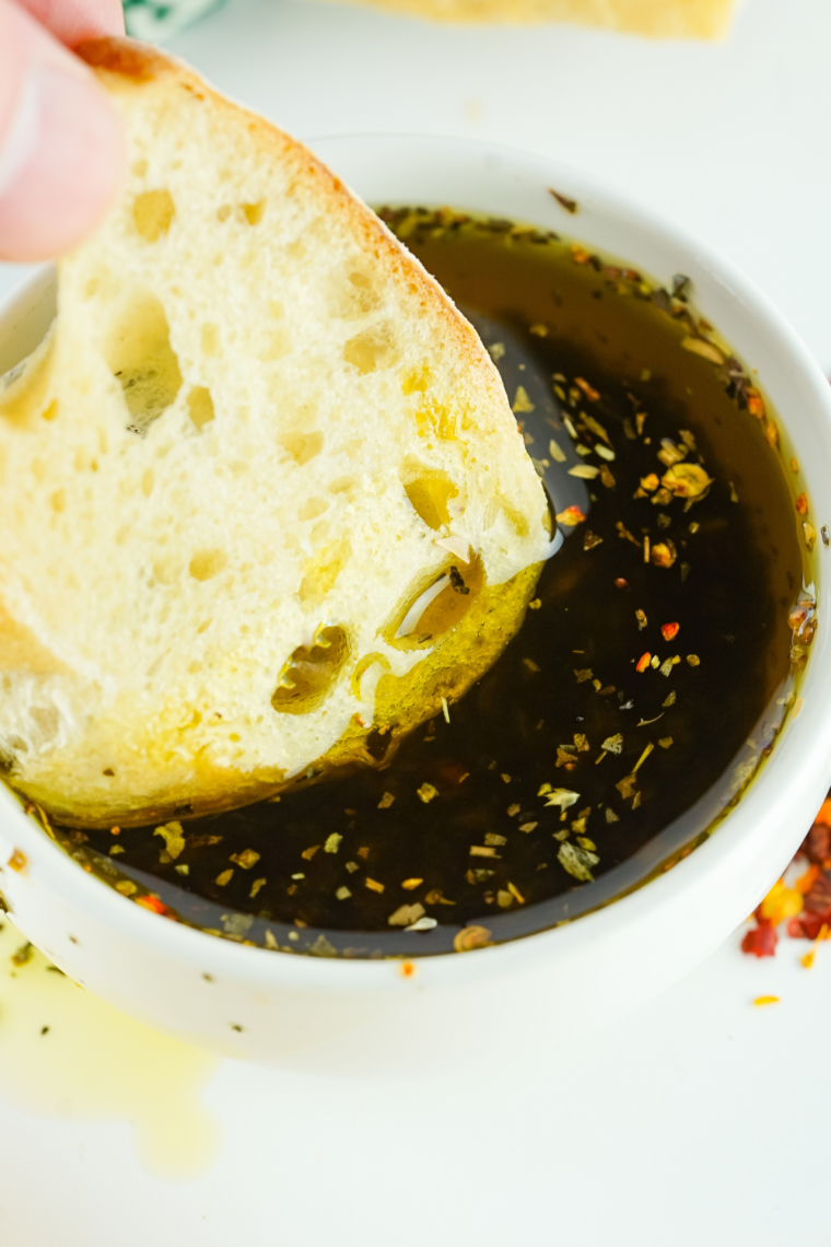 Close-up of golden bread dipping oil with fresh herbs and spices in a small bowl, served with crusty bread.