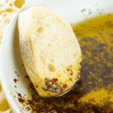 Small bowl of Greek-inspired bread dipping oil with herbs and garlic, surrounded by slices of crusty bread.