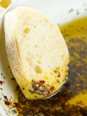 Small bowl of Greek-inspired bread dipping oil with herbs and garlic, surrounded by slices of crusty bread.