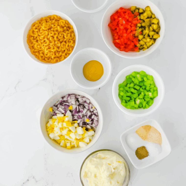 Ingredients for gluten free macaroni salad including gluten-free elbow pasta, mayonnaise, yellow mustard, gherkins, red bell pepper, celery, red onion, hard-boiled eggs, and spices.