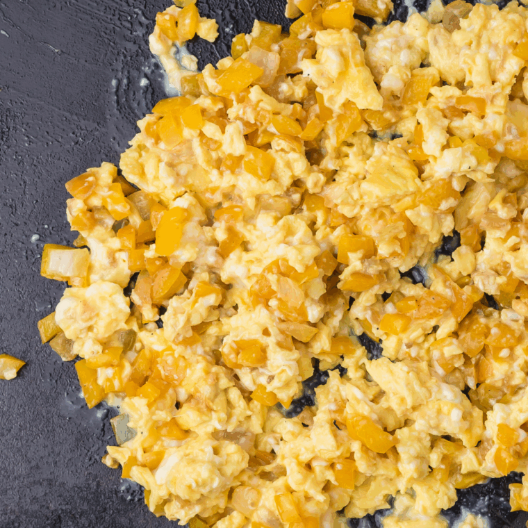 Scrambled eggs cooking beside sautéed bell peppers and onions on a Blackstone griddle