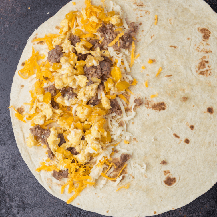 Flour tortilla topped with cheddar, mozzarella, sausage, eggs, and veggies on a griddle, ready to fold