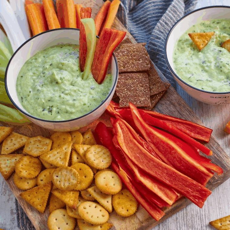 Close-up of a bowl of zucchini yogurt dip topped with fresh dill and olive oil, surrounded by fresh vegetable sticks.