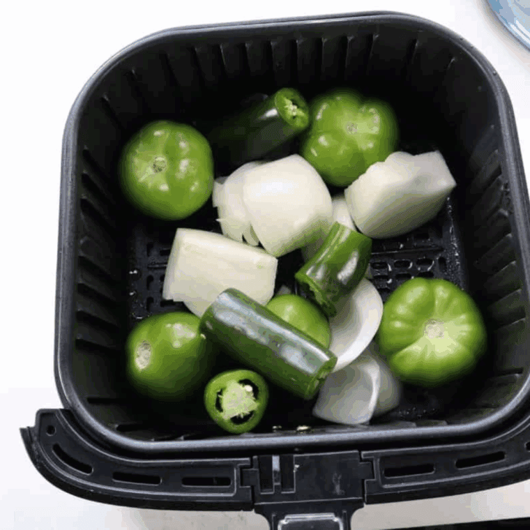 Husked tomatillos, diced onions, and garlic cooking in an air fryer basket until lightly charred and softened.
