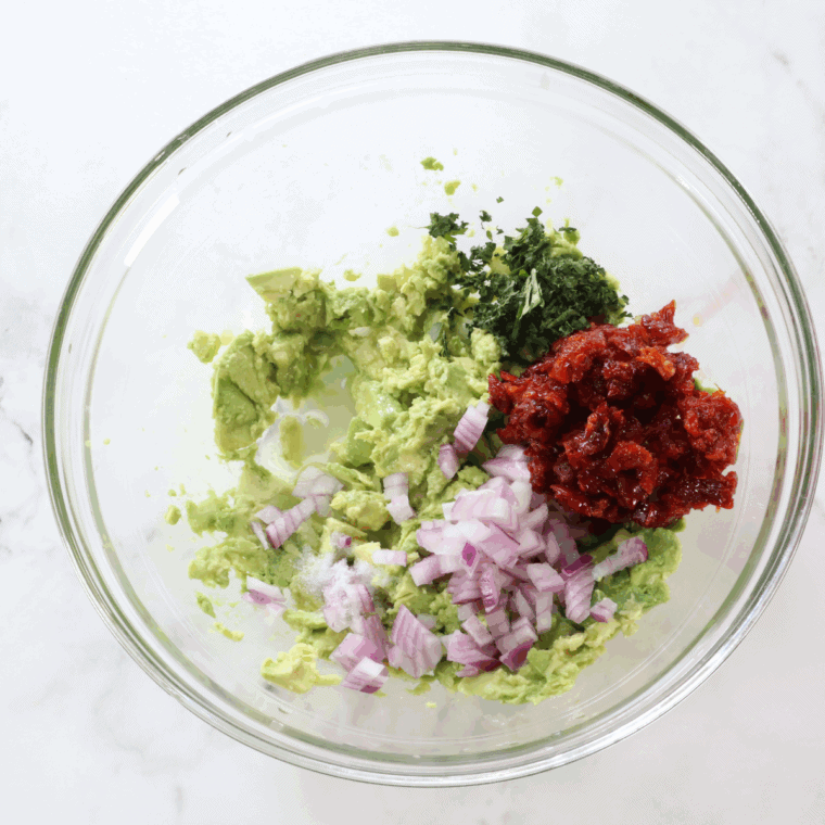 Bowl with diced avocado, sun-dried tomatoes, red onion, cilantro, lime juice, salt, and pepper being gently mixed.