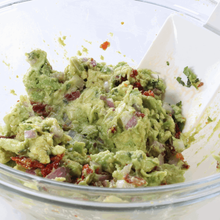Medium bowl filled with diced avocado, sun-dried tomatoes, red onion, cilantro, lime juice, salt, and pepper being gently mixed.