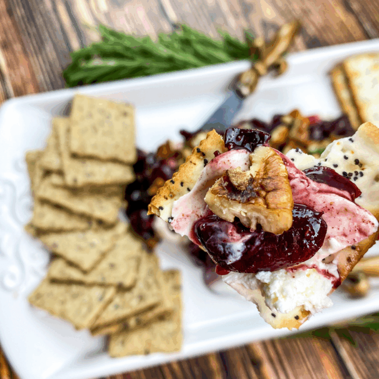 Creamy block of cream cheese topped with warm cherry sauce, pecans, and fresh rosemary, served with crackers.