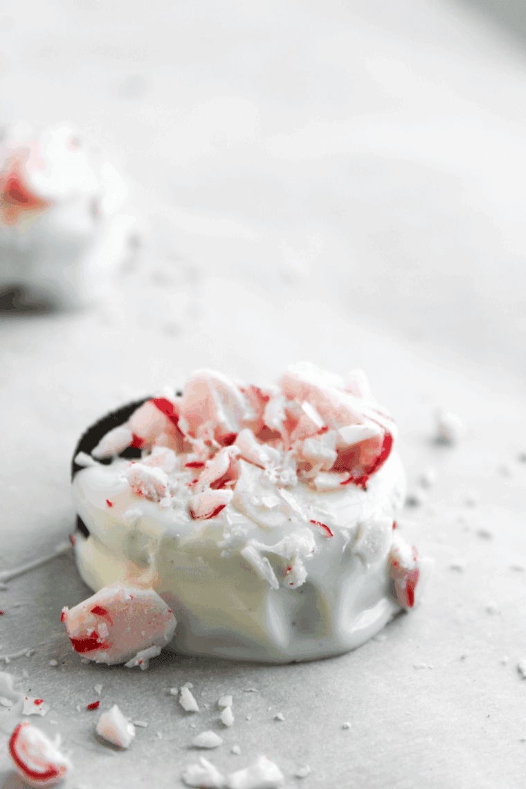 Dipping an Oreo cookie into white chocolate and immediately garnishing it with crushed peppermint on a baking sheet.