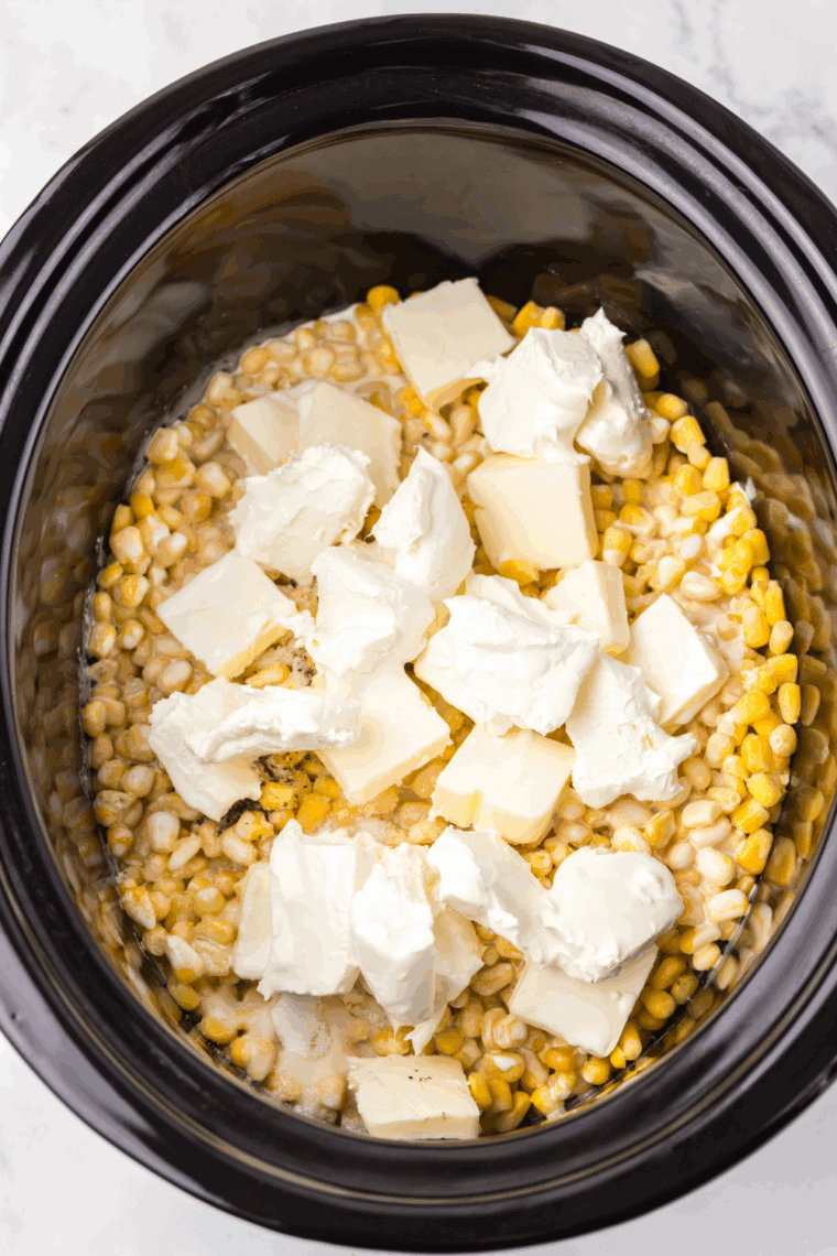 Adding corn, cubed cream cheese, and butter to a slow cooker with milk, sugar, salt, and pepper 