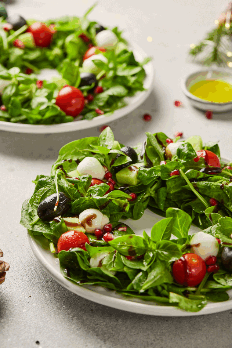 Festive Christmas wreath salad with mixed greens, cherry tomatoes, mozzarella balls, black olives, pomegranate seeds, and fresh basil, drizzled with balsamic glaze on a round platter.