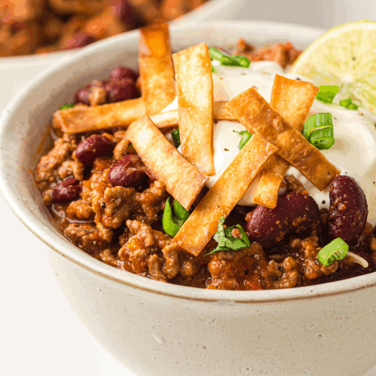 A steaming bowl of thick, beefy George’s Chili topped with a generous sprinkle of shredded cheddar cheese and finely diced red onions, served with a side of saltine crackers.