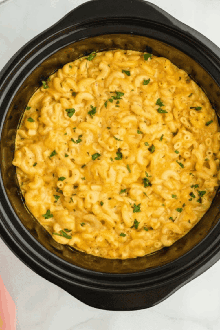 Close-up of melted cheddar and American cheese being stirred into noodles in a black slow cooker.