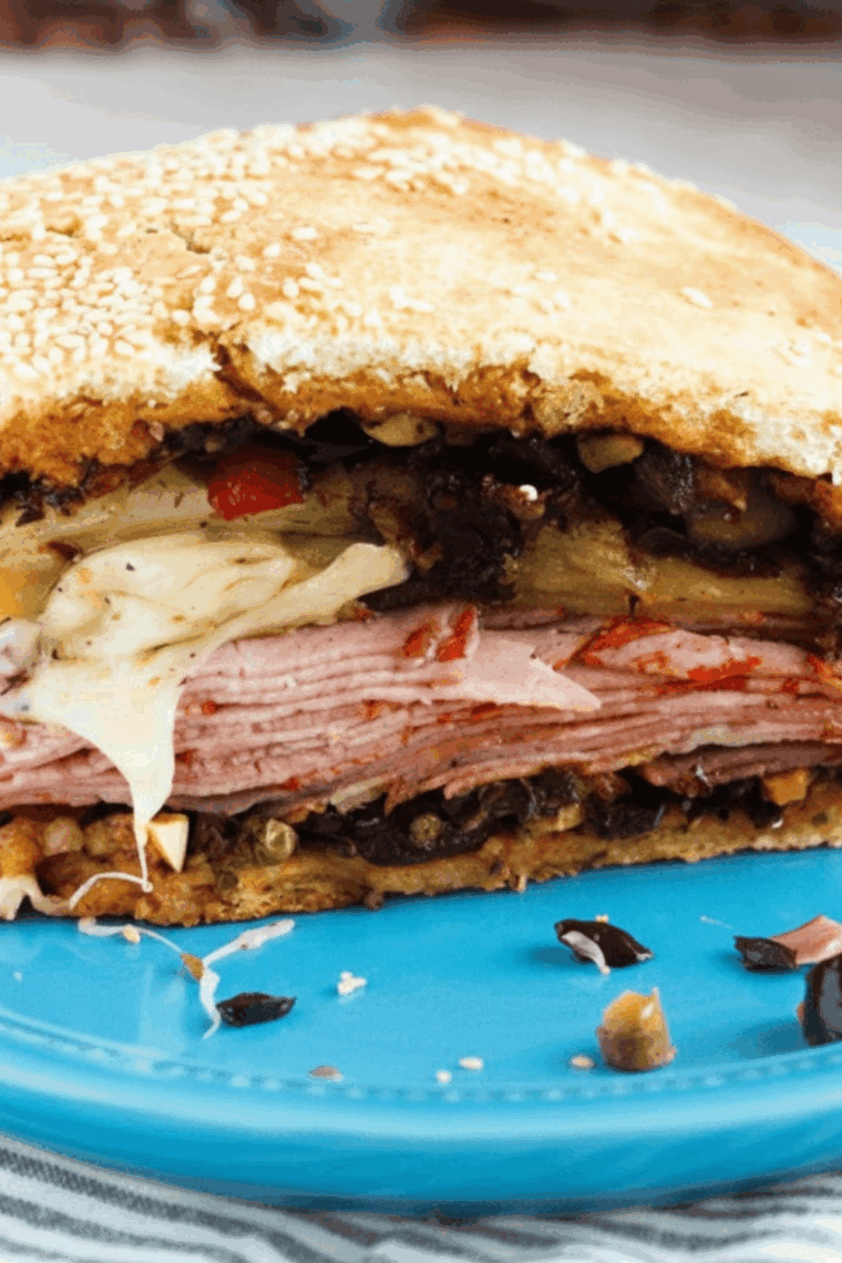 A large sliced Jason's Deli style Muffuletta sandwich showing layers of ham, salami, mortadella, and melted provolone with olive salad.