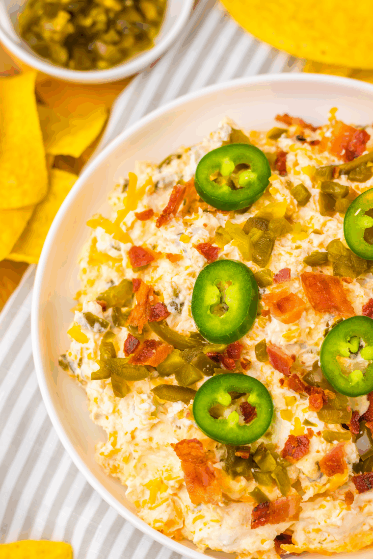 Slow cooker jalapeño popper dip topped with crispy bacon, melted cheddar, and jalapeños, served with tortilla chips
