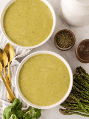 A steaming bowl of vibrant green asparagus soup garnished with fresh cream swirls and cracked black pepper.