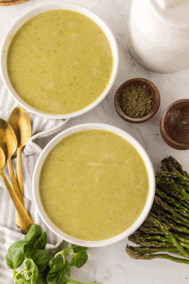 A steaming bowl of vibrant green asparagus soup garnished with fresh cream swirls and cracked black pepper.