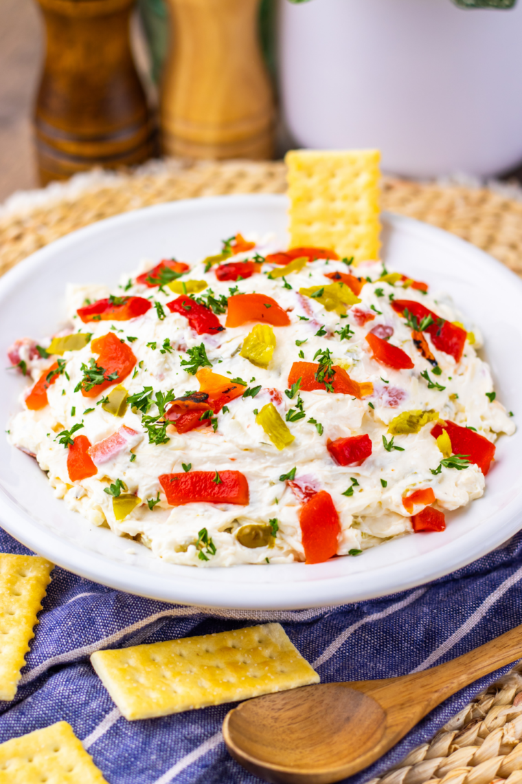 Cream cheese pepper dip served in a bowl with crackers and fresh vegetable sticks on the side.
