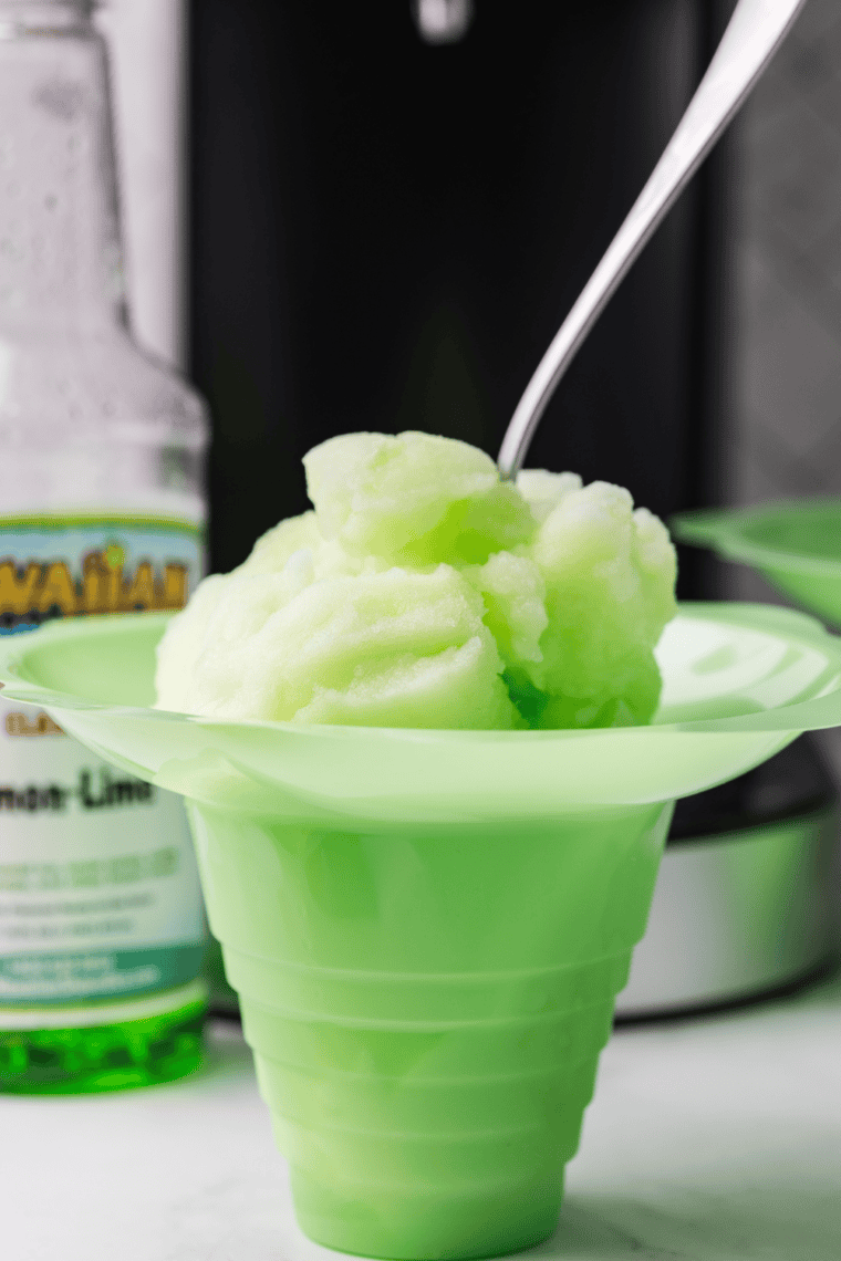 Homemade lemon-lime Italian ice in a clear glass, pale yellow-green with icy texture, garnished with a lime wedge, on a white countertop.