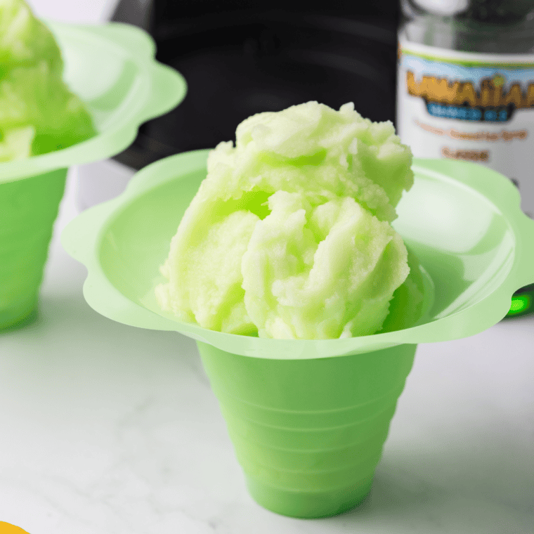 A glass of homemade lemon-lime Italian ice, pale yellow-green with icy texture, garnished with a thin lime slice, sitting on a white countertop.