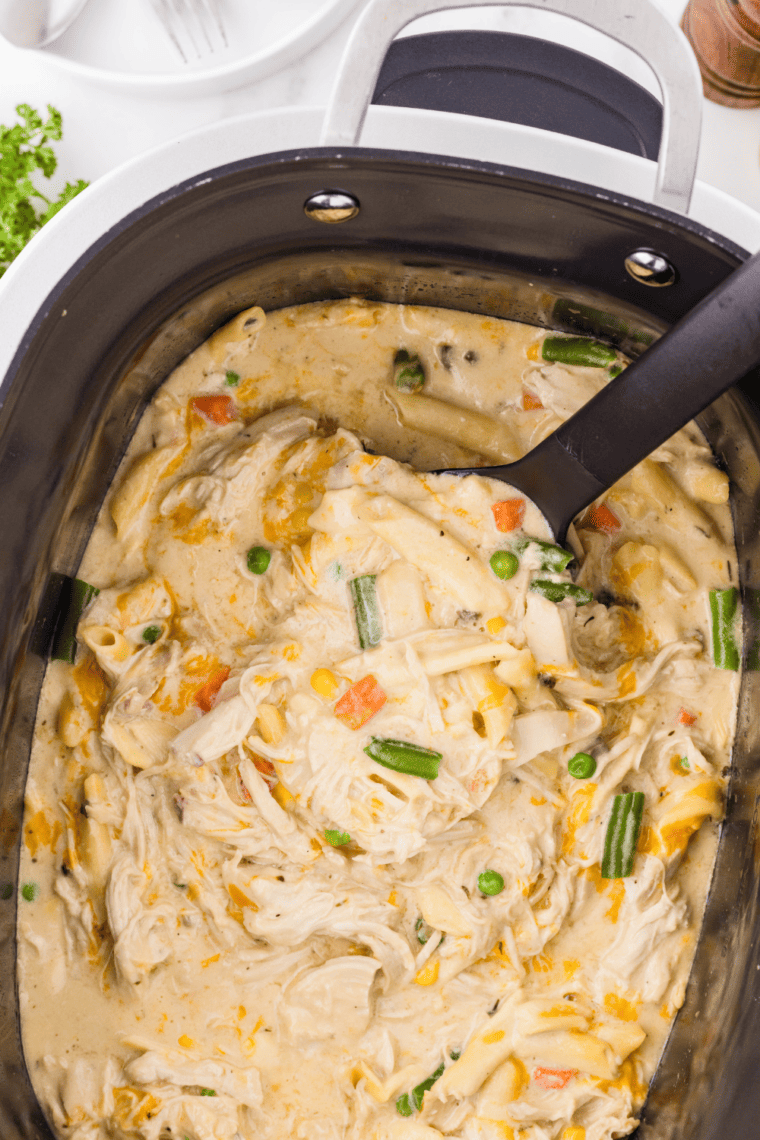 Overhead shot of creamy crock pot chicken pot pie pasta in a white bowl with shredded chicken, peas, carrots, and fresh parsley.