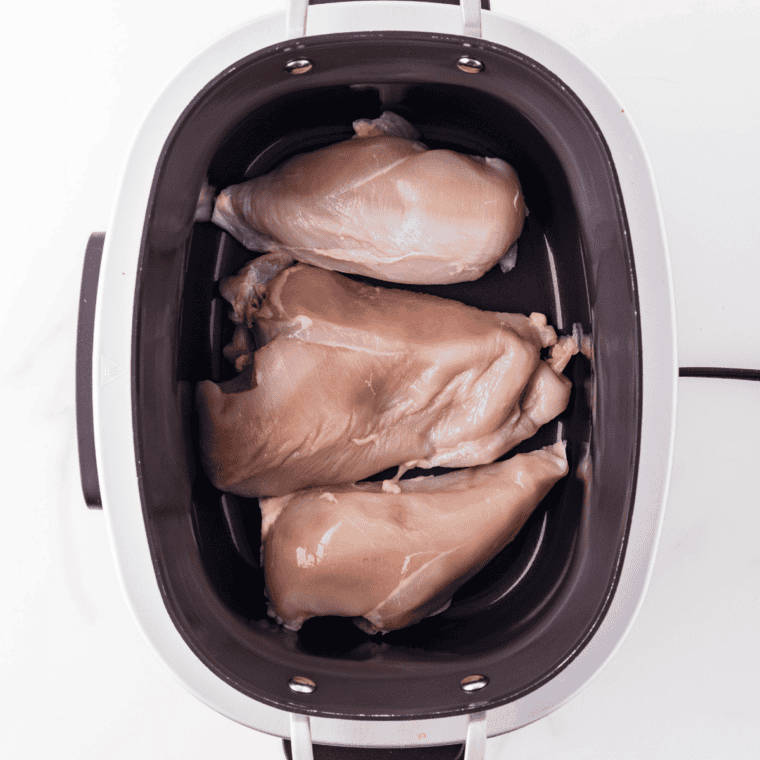 Raw chicken breasts nestled in creamy soup mixture topped with frozen mixed vegetables inside a 6-quart crock pot.