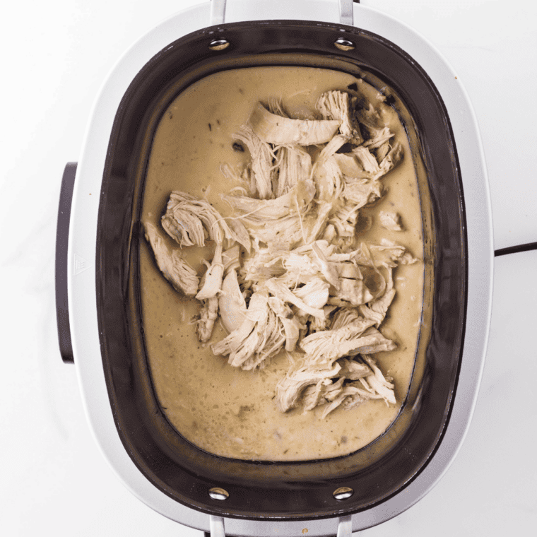 Cooked chicken breasts shredded with two forks on a wooden cutting board next to the slow cooker.