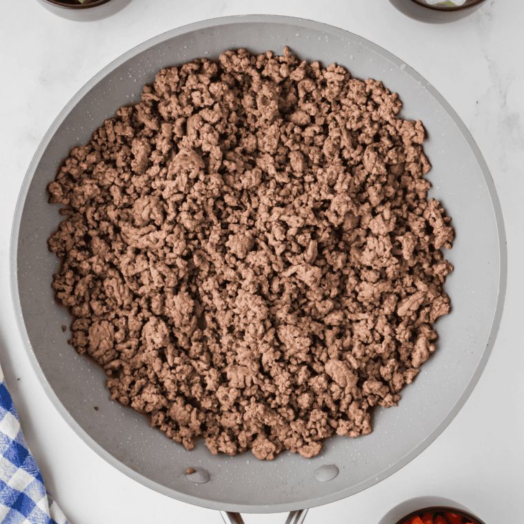 Ground beef cooking in a large skillet over medium-high heat, being broken apart with a wooden spoon, with diced onion and bell pepper added as it browns and excess grease being drained off.