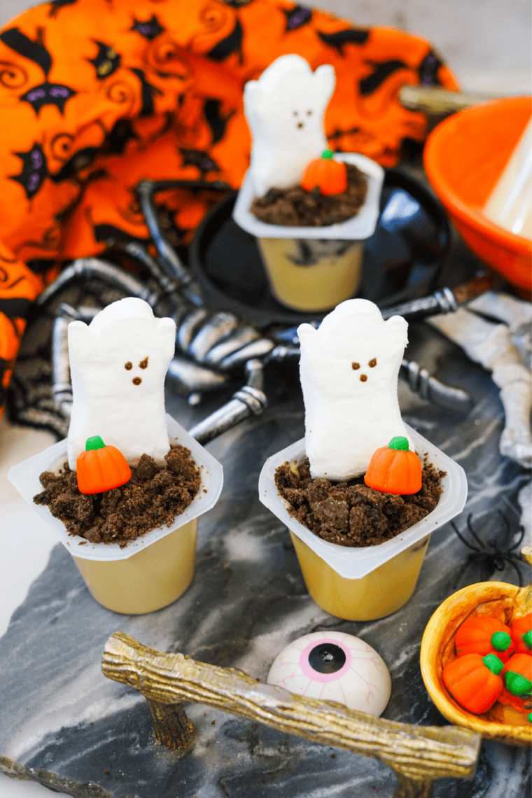 Three Halloween pudding cups with marshmallow ghosts and candy pumpkins on a dark table