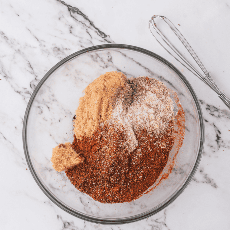 All the copycat Honey Hog BBQ rub ingredients added to a glass bowl, being whisked together into an even reddish-brown blend