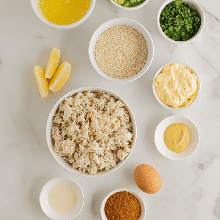 Crab cake ingredients on a kitchen table: jumbo lump crab meat, egg, mayonnaise, Dijon mustard, Worcestershire, Old Bay, panko, lemon, parsley, butter, and garlic