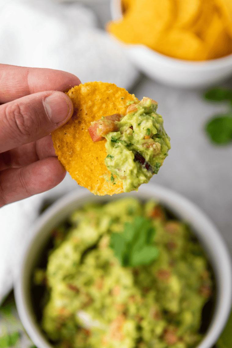 Tortilla chip scooping a generous bite of chunky spicy guacamole from the bowl