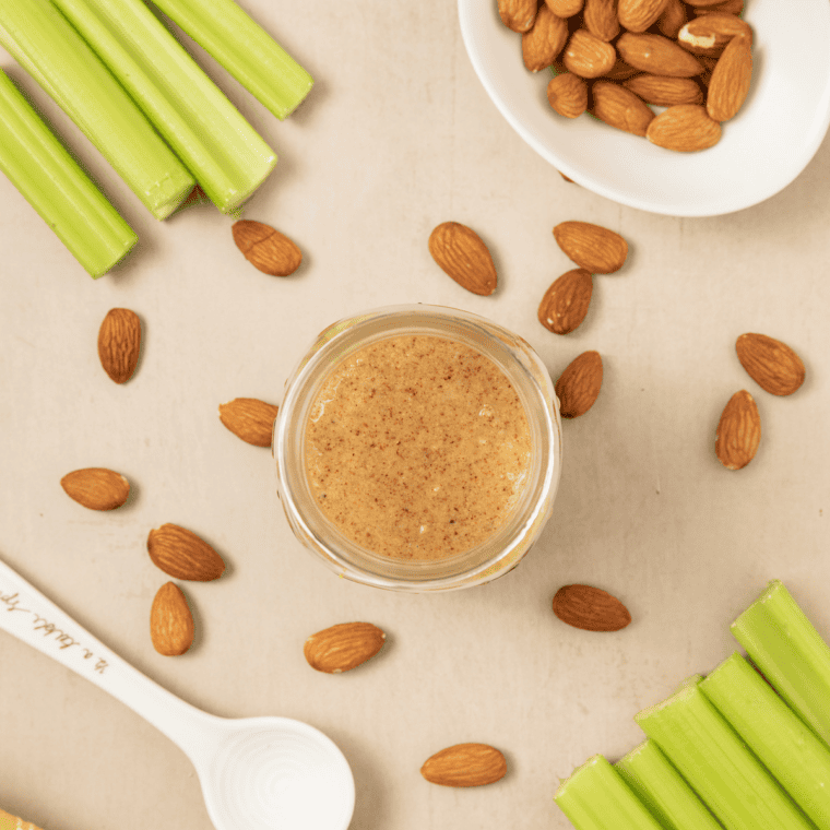 A jar of creamy almond butter surrounded by fresh celery sticks and scattered raw almonds