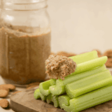 A jar of creamy almond butter surrounded by fresh celery sticks and scattered raw almonds