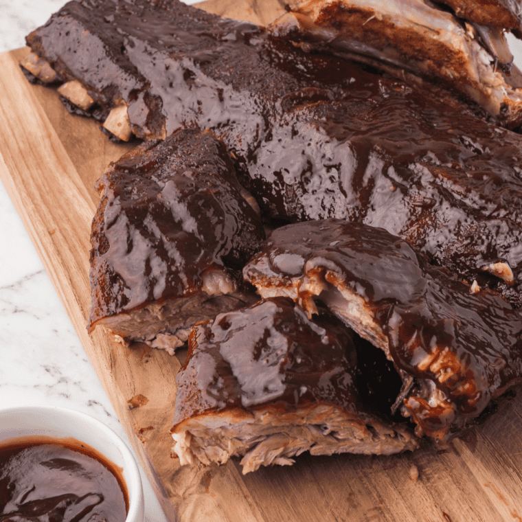 BBQ baby back ribs on a wooden cutting board beside a small bowl of barbecue sauce