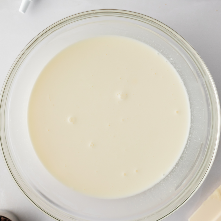 Whole milk being whisked into a cream cheese and sugar mixture until smooth and pourable.