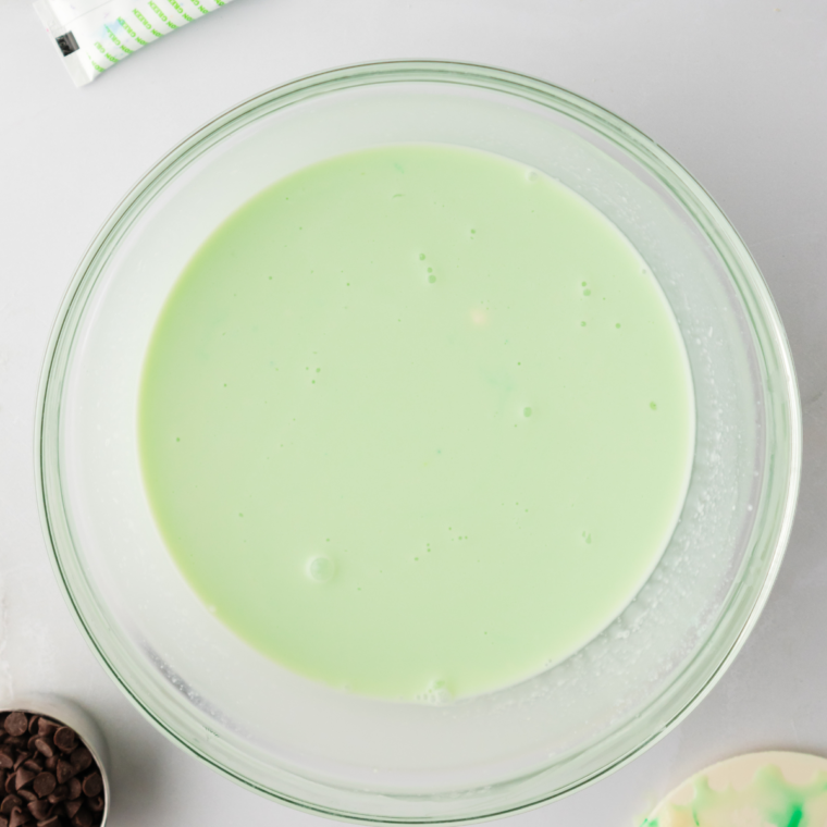 Green food coloring being added drop by drop to a bowl of mint ice cream base, tinting it bright green.