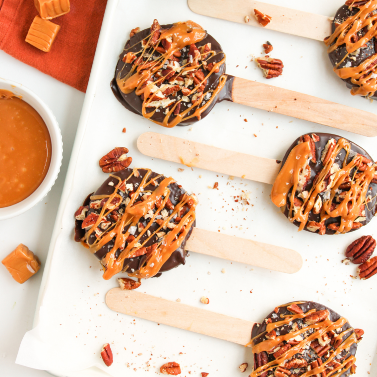 Chocolate turtle caramel apple pops on sticks, topped with chopped pecans and a caramel drizzle