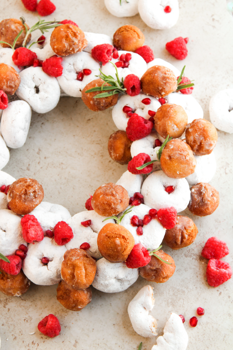 Powdered sugar donut wreath on a wooden table with rosemary and red berries tucked between the donuts