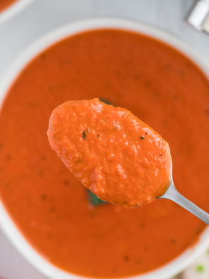 Creamy tomato soup in a white bowl topped with fresh basil and grated Parmesan, next to a grilled cheese sandwich.