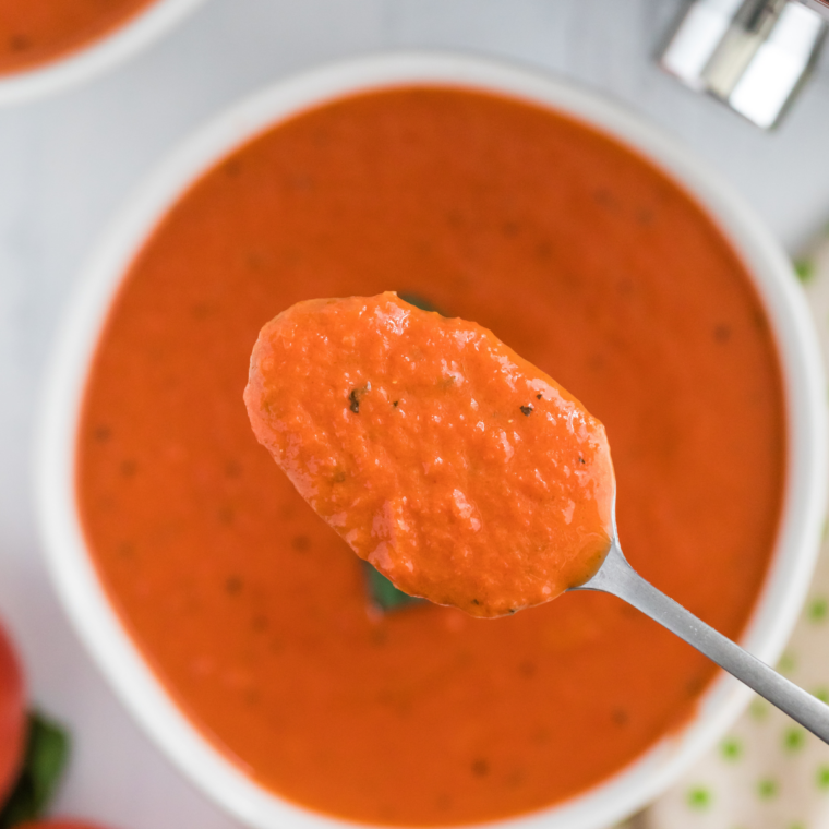 Creamy tomato soup in a white bowl topped with fresh basil and grated Parmesan, next to a grilled cheese sandwich.