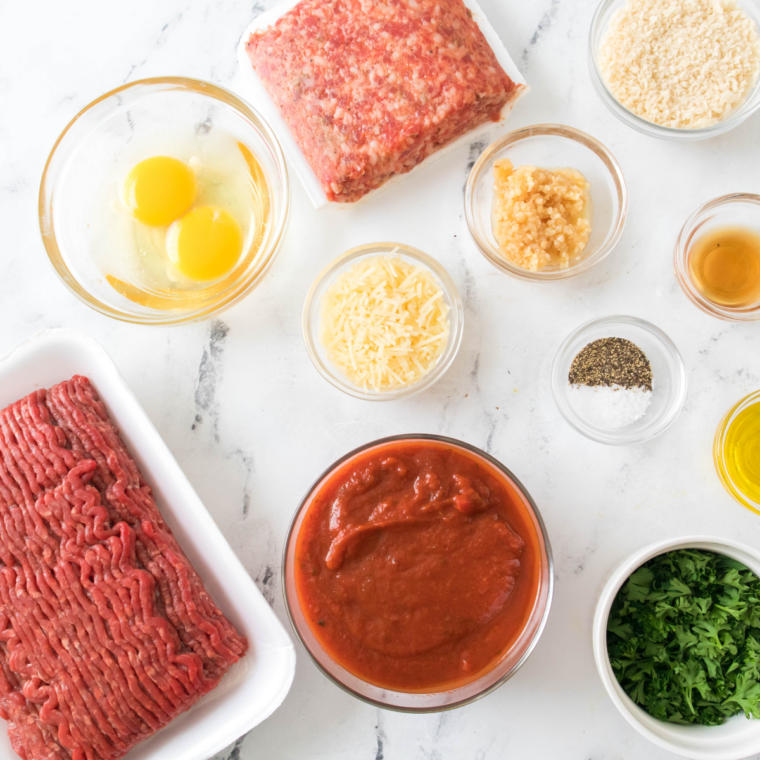 Ground beef, ground pork, panko, milk, egg, Parmesan, garlic, and parsley arranged in bowls on a marble counter