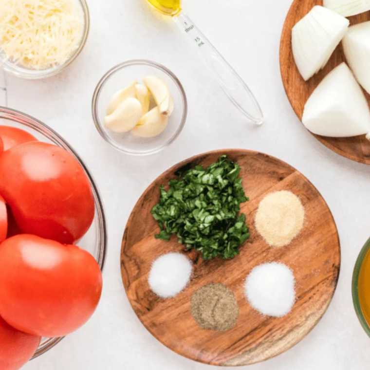 Fresh tomatoes, yellow onions, garlic cloves, olive oil and Italian seasoning on a marble counter